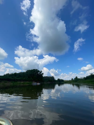 A wide, calm river flows through a lush green landscape under a vibrant blue sky filled with large, puffy white clouds. The river's surface reflects the sky and the surrounding trees, creating a mirror-like effect. On the river, a small boat with two people wearing orange life vests is visible near the left bank, which is lined with dense green foliage and trees. The scene appears peaceful and natural, likely a popular spot for outdoor recreation.