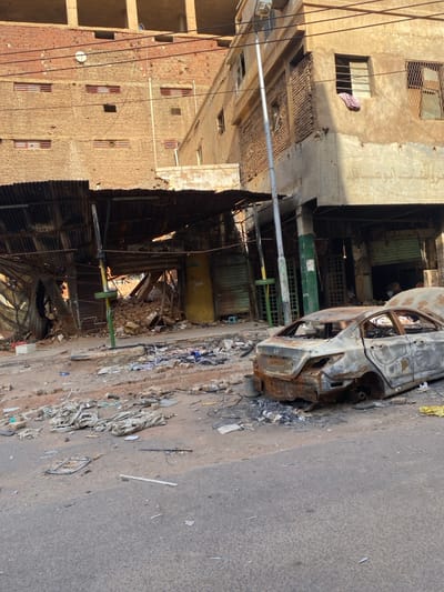 The scene shows the aftermath of a fire in a street in Khartoum, Sudan, located at coordinates (latitude: 15.602, longitude: 32.525). A burnt-out car is parked in the middle of the street, surrounded by debris. The street is narrow and flanked by two brick buildings with visible damage from the fire. The building on the right has Arabic writing on it.  A metal awning over the street is partially collapsed. The ground is covered in debris, including pieces of wood, metal, and cloth. There is no sign of human activity in the scene. The sky is not visible in the image.
