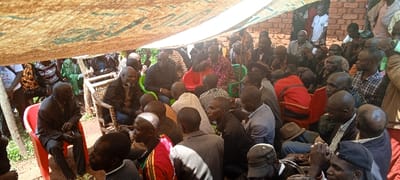 A large group of people are gathered under a tent-like structure, with many seated in plastic chairs. The attendees are diverse in age and attire, some wearing casual clothing, others in suits or traditional garb. The setting appears to be outdoors, possibly a community event or gathering. Brickwork is visible in the background behind the crowd. The lighting suggests it is daytime.
