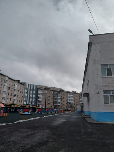 This image depicts a residential urban landscape in Nadym, Russia, under an overcast, grey sky. The perspective is looking down a pathway or street flanked by multi-story apartment buildings.
In the immediate foreground on the left, a light-colored apartment building with peeling paint and several white-framed windows is prominent. A segment of its exterior features a horizontal blue band. Adjacent to it on the right is a dark asphalt road or pathway, possibly damp, with a clear white dividing line running along its edge as it recedes into the distance.
Above, against the cloudy sky, an electrical wire stretches across the frame, from which a single street light fixture hangs. The sky is entirely covered by a thick layer of grey clouds, suggesting an overcast day.
Further into the scene, on the left side, a series of other multi-story apartment buildings rise. These buildings exhibit various architectural styles and colors, including sections with light and darker facades, and many windows. Nestled between the road and these background buildings, a children's playground is visible, featuring colorful equipment in shades of red, yellow, and blue.
No people are visible in the scene, and there is no discernible text. The overall impression is one of a typical, quiet residential area, with the cloudy weather contributing to a somewhat muted ambiance.