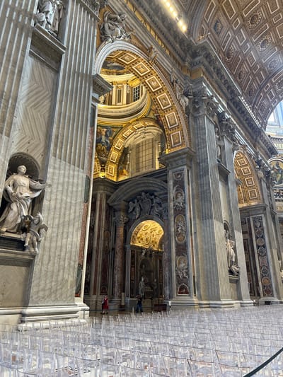 The image displays the interior of St. Peter's Basilica in Città del Vaticano, Vatican City. The perspective is from ground level, looking upwards and diagonally across the nave.
In the immediate foreground, numerous rows of empty, clear, translucent plastic chairs are uniformly arranged on a patterned floor.
To the left, a massive, light-grey fluted pilaster, constructed from marble, rises vertically. A large niche within this pilaster houses a white marble statue of a male figure, possibly a saint, depicted gesturing with an outstretched arm. Below this figure, a smaller marble putto or angel holds a cross. The lower section of the pilaster features a subtle diagonal pattern in the marble.
Moving towards the center and right, the basilica's architectural elements continue. Multiple grand fluted pilasters, identical in style and material to the one on the left, define the structure. Between these pilasters, vertical wall panels of reddish-brown marble are visible, adorned with various reliefs and frescoes depicting figures and scenes. Arches with intricate golden ornamentation and painted panels extend into the upper sections.
The vaulted ceiling is elaborately decorated with coffered squares and rectangular patterns, predominantly in gold, brown, and off-white, featuring rosettes and detailed motifs. Artificial spotlights are visible high on the ceiling structure, augmenting natural light entering through arched windows positioned in the upper architectural elements.
In the midground, near the base of the columns and arches, several individuals are present. One person wearing a red dress is walking from left to right. Another individual is partially visible further to the right. The overall lighting is a combination of natural light from windows and artificial illumination, creating distinct shadows and highlighting the detailed textures and ornamentation.