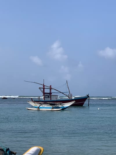 The image shows two fishing boats in the ocean off the coast of Mirissa, Sri Lanka, under a clear blue sky with scattered clouds. The larger boat, predominantly red and white, is anchored in the background, with a wooden structure and a long pole extending from it. In the foreground, a smaller, narrower boat with blue and white accents, also appears to be anchored. The water is a calm, blue-green, with gentle waves rolling in towards the shore, where rocks are visible in the distance. The scene is peaceful and depicts a typical coastal fishing environment.