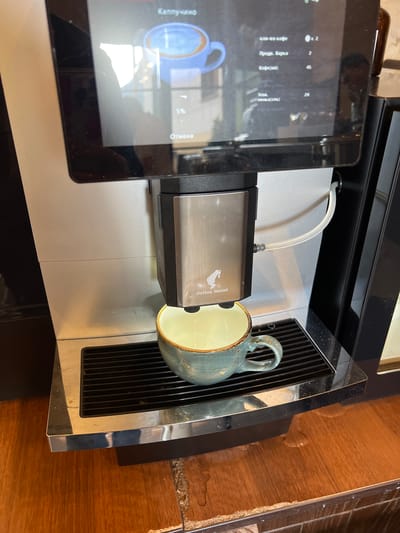 The image shows a close-up, eye-level view of a cappuccino being made in a coffee machine. The coffee machine has a digital display with Russian text, indicating options for making coffee. A light blue cup with a frothy top is visible on the display screen. Below the screen, a chrome nozzle is dispensing a liquid, possibly milk or coffee, into a turquoise ceramic mug with a textured finish. The mug is placed on a black drip tray with horizontal slats. The coffee machine itself is a brushed metal and black appliance, with a transparent tube exiting from the right side. The background is blurry, but appears to be a wooden countertop or table. The lighting suggests an indoor setting. There is no indication of the location being Dombay, Russia.