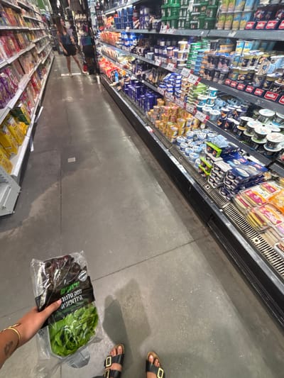 This image depicts an interior view of a supermarket aisle within a Shufersal branch located in Bnei Dror, Israel. The perspective is from an individual looking forward, holding a product.

In the immediate foreground, a person's left hand, adorned with a thin gold bracelet, grasps a clear plastic bag containing 'Shufersal Hydroponic Salanova Lettuce' (חסה סלנובה הידרופונית), a mix of green and red lettuce leaves. The person's feet are visible, wearing black sandals with gold-colored buckles, standing on a grey concrete floor that exhibits minor cracks and scattered light-colored specks.

To the left, a tall shelving unit displays various packaged dry goods, including cereals and snacks, extending into the background. To the right, a long refrigerated display case holds multiple tiers of dairy products such as yogurts, puddings, and cheeses, alongside pre-packaged deli items. Further down the aisle, in the mid-ground and slightly to the left, a blurred female figure is visible, wearing a dark top and skirt or shorts, with light-colored footwear. The scene is illuminated by overhead artificial lighting typical of a retail environment.
