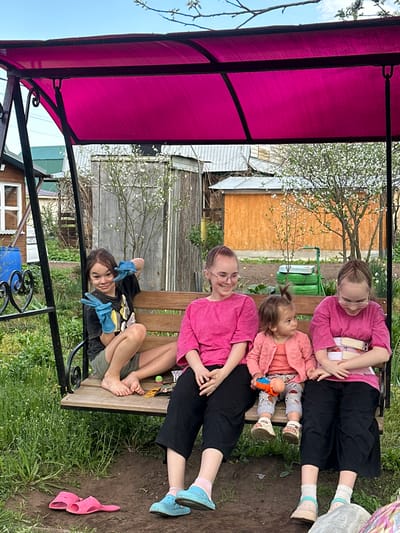 The image shows a group of four people sitting on a wooden swing in a backyard setting. The swing is covered by a pink canopy. Three individuals, including a woman, a girl, and a toddler, sit together on one side of the swing, all wearing pink tops. The woman wears glasses. The toddler holds a small doll. On the other side of the swing, a younger individual wearing blue gloves poses with hands raised. The environment includes a house, a wooden fence, green grass, and trees with white blossoms. A pair of pink sandals lies on the ground. The location is possibly Uva, Russia.