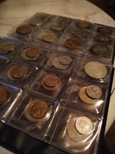 The image shows a close-up, overhead view of a coin collection album. The album is black and appears to be made of plastic or leather. It contains several clear plastic pages, each with multiple rectangular slots. Each slot holds one or more coins. The coins vary in size, color, and apparent denomination. Some slots contain a single coin, while others hold two coins stacked on top of each other. The lighting is dim and appears to be coming from above and to the right, creating highlights and shadows on the coins. The background is blurred but suggests a textured surface, possibly a tablecloth or a desk. There are no people or animals visible in the image, and no indication of a specific location within Венюково, Russia, or any other geographical area. The scene is static and focused on the collection of coins.