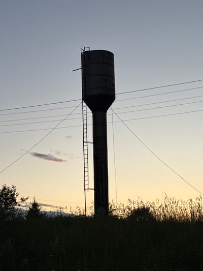A silhouetted water tower stands tall against a pale, hazy sky. The tower, a cylindrical structure with a slightly wider top, is equipped with a ladder that runs vertically up its side. Several power lines stretch horizontally across the sky, intersecting the tower at different heights. In the foreground, tall, dark grass and vegetation create a dense, shadowed border, obscuring the ground. A small, elongated cloud with a pinkish hue is visible in the distance, adding a subtle touch of color to the otherwise muted sky. The scene appears to be set in a rural or semi-rural area, possibly on the outskirts of a town or village in Kukui, Russia, given the presence of the water tower and the natural landscape. The overall impression is one of quiet solitude and the looming presence of industrial infrastructure against a fading sky.