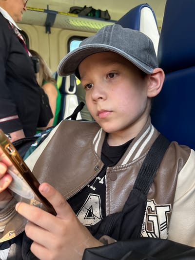The image depicts a young male passenger within the cabin of what appears to be public transport, such as a train or bus, located in Vyatskiye Polyany, Russia. The boy, positioned in the foreground, wears a gray flat cap and a brown and off-white faux leather varsity jacket featuring striped ribbed cuffs and collar. Beneath the jacket, a black t-shirt with a distressed white "A" graphic is visible, along with an embroidered "GT" patch on the right sleeve. A black cross-body bag strap is slung over his right shoulder. He is looking downwards, absorbed by a dark-colored smartphone held in his right hand, which also secures a small, semi-transparent white object, possibly a card or ticket. He is seated in a blue upholstered seat with a white headrest element. In the mid-ground to the left, an adult, potentially a female, is partially visible, wearing a dark jacket over a white collared shirt and a red and white striped tie, indicative of a uniform. She wears glasses. Further back, another passenger wearing a bright green top is partially obscured. The cabin interior features light-colored walls and an overhead luggage rack with a mesh base and yellow labeling. Natural light enters through the windows, revealing a bright outdoor scene with green foliage.