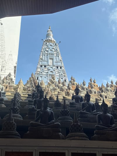 The image captures a low-angle view of the Gangaramaya Temple in Colombo, Sri Lanka. The temple's distinctive silver-colored pagoda rises toward a bright blue sky with light clouds. In the foreground, rows of seated Buddha statues line the temple walls. The statues appear in various shades of gray and tan, possibly due to lighting and material differences. A portion of a building with a dark roof and a white wall are visible on the left side of the frame. The photograph is taken from a position that looks up toward the temple and sky, creating a sense of height and grandeur.