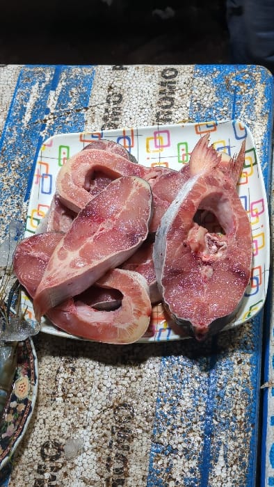 Slices of raw fish steaks are piled on a patterned rectangular plate. The fish is pink with a visible bone structure in the center of each steak. The plate sits on a worn, blue-and-white patterned surface, possibly a cooler or container. The surface appears to be made of styrofoam-like material. Part of what appears to be some shrimp is visible in the bottom corner of the image.