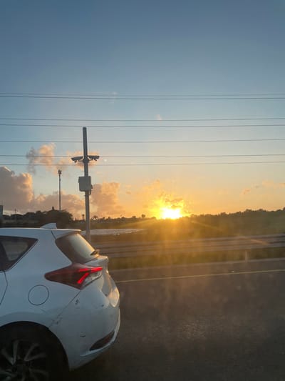 A white car is parked on the side of a road at sunset. The sun is low in the sky, shining through clouds near what appears to be a line of trees. There are utility poles and lines visible. The car's taillights are illuminated, and there is visible damage to the rear of the vehicle. Hod HaSharon, Israel, cannot be definitively confirmed with the information present.