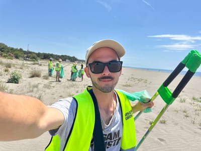 A selfie shows a man wearing sunglasses, a light-colored baseball cap, and a neon yellow safety vest over a grey t-shirt with black text, standing on a beach in Donji Štoj, Montenegro. He is holding a green and black trash picker in his left hand. A group of people wearing similar safety vests are picking up trash in the background. The beach has sand, some vegetation, and in the distance, some trees. The sky is blue. The image is rotated approximately 180 degrees.