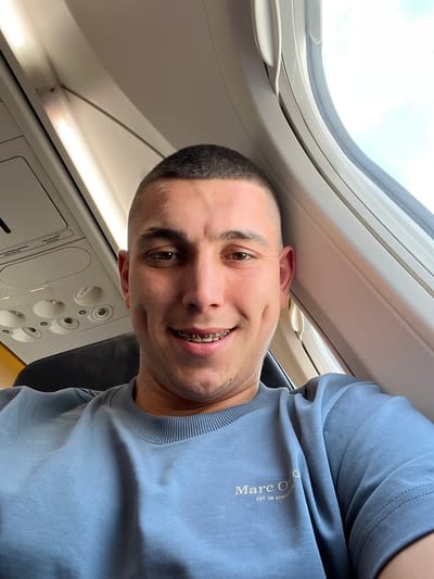 A man is seated in an airplane, next to the window. He is wearing a blue T-shirt with the words "Marc O'Polo EST. IN STOCKHOLM" printed on it. He has short, dark hair and is wearing braces. The window shows a view of the sky. The interior of the airplane cabin is visible, including the overhead panel with buttons and vents. The location, Eindhoven, Netherlands, cannot be determined from the image.