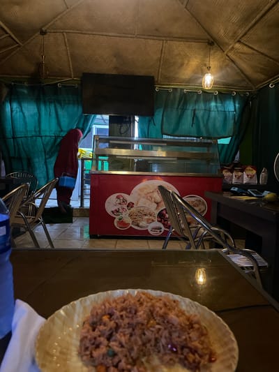 The image depicts an indoor dining area in what appears to be a restaurant in Hayin Banki, Nigeria. The scene is partially obscured by a plate of rice-based food in the foreground. There is a person with a red shirt near a refrigerator with food items on its side in the back. Green fabric covers some of the surroundings. Several metal chairs are visible. Lighting fixtures are present on the wall. The structure of the building is visible on the left side.