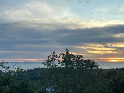 The image shows a scenic view at sunset in Baan Hin Kong, Thailand. The horizon features a body of water, likely the sea, with a scattering of islands in the distance. The sun is setting on the right side of the frame, casting a golden and orange glow across the water and sky. The sky is filled with layers of clouds, ranging in color from gray and blue to yellow and orange. A bird is visible in the sky. In the foreground, there are lush green trees. A building with a light-colored roof, potentially a residence, is partially visible among the trees. The scene appears peaceful and natural.