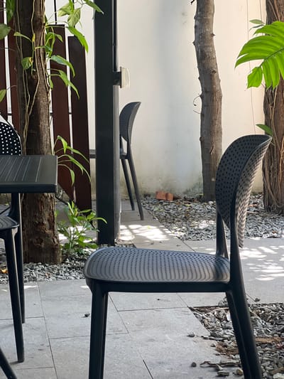 This is an outdoor scene in Siem Reap, Cambodia, likely a cafe or restaurant patio. In the foreground, a black plastic chair with a perforated seat and back is angled towards the viewer. To its left, a corner of a black table with a perforated surface is visible, supported by slender legs. A small green plant grows in a patch of grey gravel near the table's legs.
Behind the foreground chair, a thin, dark vertical post, part of a structure, stands. Further back, another black plastic chair is positioned, its form visible through the post. A narrow pathway of grey gravel is visible between the foreground and the background wall, which is painted white. Trees with rough bark stand behind the chairs, and a large, green leaf from one of these trees is visible in the upper right corner. Vines with smaller leaves climb one of the trees on the left, adjacent to a dark wooden fence. The ground is paved with square grey tiles. The overall lighting suggests it is daytime, with dappled shadows cast by unseen foliage.