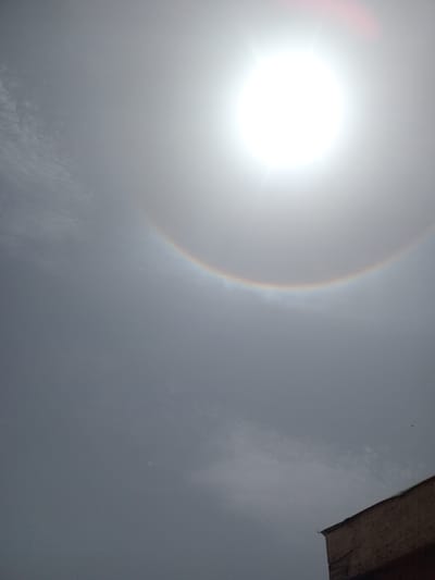 The image displays a bright, overexposed sun positioned in the upper left quadrant of the frame, surrounded by a prominent solar halo (22-degree halo). The halo, a meteorological phenomenon caused by ice crystals in the atmosphere, exhibits subtle iridescent colors, forming a distinct arc around the sun. The sky is predominantly a hazy or overcast gray, with faint textural variations indicating cloud cover. In the upper right corner, a dark, angled edge of a wooden structure, possibly a roof or eave, is partially visible. The scene captures an outdoor daytime event, likely around midday or early afternoon given the sun's high position and brightness. The weather conditions indicate the presence of high-altitude cirrus clouds necessary for the halo formation, combined with lower-level atmospheric haze. There is no visible text in the image. The location context for this media file is Agingi Zion, Nigeria.