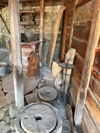 The image depicts the interior of a rustic wooden structure, likely a historical exhibit or outbuilding, situated in the town of Zheravna, Bulgaria. The floor is paved with irregular flat stones, and the walls are constructed from rough-hewn stone. Dominating the foreground are two large, circular millstones, one partially visible in the lower center and another positioned behind it. To the left, a weathered wooden post supports the structure, with a metal bucket hanging from a chain nearby. Further back, a carved wooden bear sculpture sits on the stone floor. Various other wooden elements, including beams, planks, and a wooden structure with chains and gears, are visible throughout the space, suggesting it may have been a workshop or storage area. A wrought-iron candle holder stands on the right side of the image, leaning against the wooden wall. The overall impression is one of age and tradition.