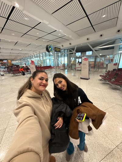 Two smiling women are captured in a selfie within an airport terminal in Nouaceur, Morocco. The woman in the foreground wears a light beige hoodie, while the woman behind her is dressed in a black long-sleeved top (possibly with "LA" visible text), blue jeans, and brown boots lined with white faux fur. She holds a smartphone with a distinctive neon green case.

They are seated on the tiled floor of what appears to be a waiting area, with rows of empty red airport chairs visible in the background. The terminal features a modern design with a ceiling composed of white panels and integrated lighting. Various airport infrastructure elements are visible, including glass walls, support beams, and informational displays.

A large circular clock to the left of the women shows a time of approximately 10:15 or 10:20 and displays "F2 F1," likely indicating gate numbers. Further in the background, a prominent white sign with red accents clearly reads "PRIORITY BOARDING." Other digital screens and stand-up signs are present but illegible. Both women are looking directly at the camera with happy and relaxed expressions, suggesting a moment of leisure before or after travel.