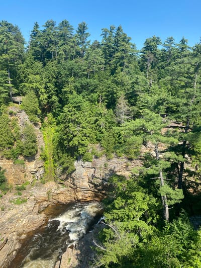 The image depicts a natural landscape with a waterfall cascading down a rocky cliff face, surrounded by lush green coniferous trees. The water is turbulent and foamy as it descends. The rocky cliff shows layers of geological strata, with patches of green vegetation growing from its crevices. Several people are visible on the cliff face, appearing to be observing or interacting with the natural scenery. Some are positioned on ledges, while others seem to be walking or climbing. In the lower left portion of the image, there is a wooden structure, possibly a small building or part of a viewing platform, nestled amongst the trees and rocks. The sky is clear and blue, suggesting a sunny day. The overall scene conveys a sense of natural beauty and outdoor recreation. The location is identified as the Town of Peru, USA.