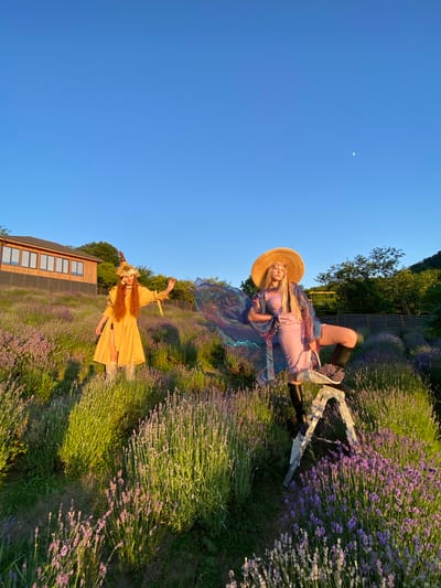 In a lavender field under a clear blue sky with a sliver of moon visible, two women are featured. The woman in the foreground, standing in a bright yellow dress and a floral headdress, has her arms outstretched. She is in a lavender field with a wooden building in the background. The second woman, seated in a wagon amidst the lavender, wears a straw hat and a sheer, iridescent shawl over a pink dress. She has a basket filled with lavender in the wagon. The scene appears to be during the golden hour, with the sun casting a warm glow over the landscape. The location is Verkhneye Uchdere, Russia.