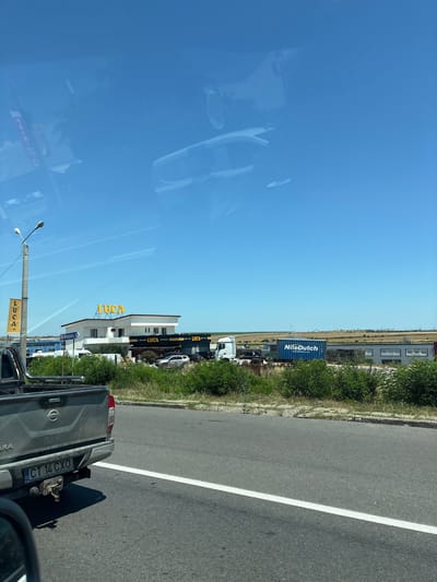 The image depicts a street scene in Lazu, Romania, with a clear blue sky overhead and scattered white contrails. In the foreground, the rear of a grey Nissan pickup truck is visible, with its license plate reading "CT 14 CXO". To the left of the truck, a street light pole with a yellow banner displaying "LUCA" is present.

Further down the road, a modern building with white and dark accents is visible, featuring large "LUCA" signs in yellow lettering on its roof and facade. Several cars are parked in front of the building. Adjacent to this building, a white truck and a blue shipping container with "NileDutch" branding are parked. The background shows a flat, rural landscape with distant structures and vegetation. The overall scene suggests a commercial area on the outskirts of a town.