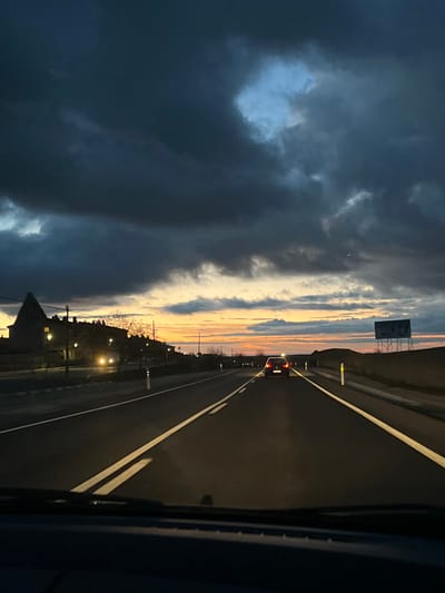 The image depicts a twilight scene from the interior of a vehicle traveling on a two-lane asphalt road in Palazuelos de Eresma, Spain. The sky is largely covered by dark, heavy clouds, with a band of orange, yellow, and light blue hues visible at the western horizon, indicative of a recent sunset.

The road ahead features a double solid white line marking the center, with a single solid white line delineating the right shoulder. A dark-colored sedan, likely a Volkswagen Golf, proceeds ahead in the same lane, its red taillights illuminated. On the left side of the road, several buildings are visible, including a prominent structure with a dark, pointed roof and illuminated windows. Multiple streetlights cast a yellowish-white glow on the roadside, and at least two sets of bright white headlights from oncoming vehicles are discernible in the distance. Bare trees or shrubs are present alongside the road and near the buildings.

The right side of the road displays undulating, sparsely vegetated terrain, with a large, empty metal billboard structure standing on tall poles further down the road. White reflective road markers line both shoulders. The bottom portion of the image shows the dark dashboard and windshield frame of the viewer's vehicle.