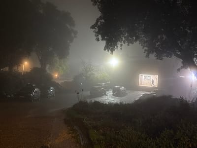 This nighttime photograph captures a heavily fog-laden scene, likely in a rural or semi-residential area within the Gilboa Regional Council, Israel. The pervasive fog diffuses all light sources, creating halos and a soft, mysterious glow that permeates the entire image.
In the immediate foreground, an unpaved, earthy path leads into the scene, transitioning to a paved area where several vehicles are parked. On the left, a dark-colored SUV is clearly visible, with a smaller car partially obscured behind it. Large, dark trees with dense foliage frame the left and right sides of the image, their canopies silhouetted against the hazy backdrop. Further back, more trees and dense vegetation are visible through the mist.
Various artificial lights pierce the fog. A warm, orange streetlamp glows on the far left, partially hidden by foliage. A brighter, whiter light source is visible on the far right, possibly another street light or building light. The most prominent illumination emanates from a brightly lit doorway of a simple building located on the right side of the frame. Within this illuminated rectangular opening, a solitary human figure is silhouetted, appearing to stand or walk, adding a sense of scale and human presence to the otherwise quiet scene.
The heavy fog creates a dreamlike and somewhat isolated ambiance, emphasizing the glow of the lights and obscuring distant details. The scene conveys a profound sense of quietude and mystery on this misty night. No discernible text is visible on any objects.