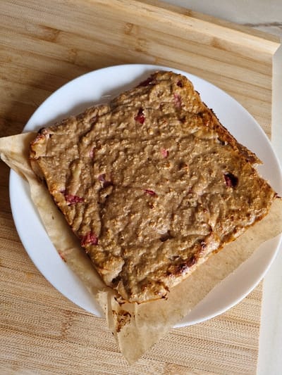 An overhead, static image depicts a single, roughly rectangular baked item situated on a white, circular plate. The baked item presents an uneven, light brown surface texture with embedded irregular reddish-pink fragments, consistent with fruit such as berries. The perimeter of the item displays a darker, caramelized brown hue. It rests on a piece of light brown, crinkled parchment paper, which extends beyond the item's base and partially covers the plate. The white, unmarked plate rests on a light-toned wooden surface, characterized by distinct horizontal grain patterns. No individuals or active events are depicted. The scene is located indoors in Niedernhausen, Germany.