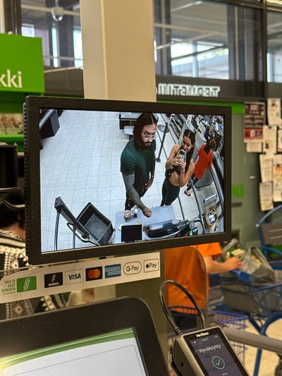 The image depicts an interior scene within an S Group supermarket located in Imatra, Finland. The central focus is a black framed monitor displaying a security camera feed of a self-checkout station. On the monitor, a male individual with dark hair, a beard, glasses, and prominent arm tattoos, dressed in a dark green t-shirt, is seen interacting with the self-checkout machine. Adjacent to him, a female individual with dark hair, wearing a black sports bra and dark leggings, holds a smartphone, seemingly recording. A black shopping basket rests on its side next to the self-checkout. Further in the background of the monitor feed, a person in an orange top is pushing a shopping cart.
In the foreground, below the monitor, a Verifone payment terminal displays "Hyväksytty" (Accepted) with a green checkmark, indicating a successful transaction. A white sticker affixed to the counter lists accepted payment methods: S-Bonus, S Business, Visa, Mastercard, American Express, and G Pay, alongside the text "MEILTÄ SAAT BONUSTA s-kanava.fi" (We give you bonus s-kanava.fi). The overall environment includes a light-colored tiled floor, green store signage partially visible in the upper left, and a reflection of the reversed word "TOIMETSA" on a glass surface in the upper background, indicating a large indoor space with visible exterior windows. A section of a blue shopping cart is visible on the right side of the image.