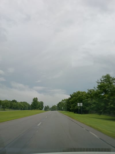 This media file shows a roadway in Lexington, United States. The perspective is from a vehicle moving along the road, which is paved and has dashed white lines marking the lanes. On the left side of the road, there is a grassy embankment with trees. In the distance, there are two road signs. The sign on the right reads "27 SOUTH" and "27 WEST" with smaller signs below that read "27 NORTH" and "27 WEST." The sign on the left reads "27 SOUTH." The sky is overcast with clouds, suggesting a cloudy or possibly rainy day. The time of day is not clearly discernible due to the cloud cover. The scene appears to be a rural or suburban area. There is no activity or event taking place other than driving. No people are visible in the media file.