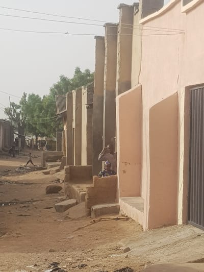A daytime shot in Dutsinma B, Nigeria, showcases a narrow street lined with buildings. The structures are constructed from light brown materials, possibly mud or adobe brick. The camera angle, positioned from a low point and tilted, captures the underside of the buildings' overhangs, which create a rhythmic pattern of projecting ledges.
A figure, presumably a resident, is partially visible under the overhang. The person is wearing what appears to be a patterned shirt.
Overhead power lines are visible, and a few trees with green foliage are seen. The ground is uneven and covered with debris, indicative of an unpaved street. The lighting is diffused, suggesting an overcast day.