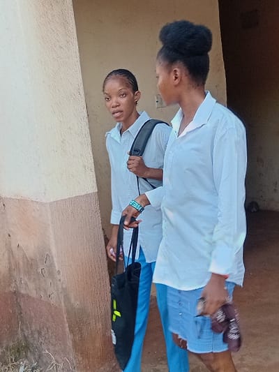 Two young women stand side-by-side. The woman in front wears a white shirt, blue jeans, and carries a black bag. The second woman, slightly behind, also wears a white shirt and jeans. Both have dark hair and appear to be outside, near a building with a neutral-colored exterior. The first woman is closer to the building's wall. The scene is well-lit, capturing the details of their clothing and surrounding environment.
