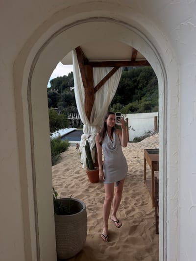 A woman is taking a selfie in a mirror. She is standing on sand, likely on a beach. She is wearing a silver, sparkly, halter dress and black flip-flops. The background shows a white wall, some greenery, and a wooden structure with white curtains. The scene appears to be outdoors in a relaxed setting. The location is identified as Drobnići, Montenegro.