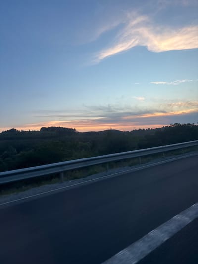 The image depicts a road scene with a view of a forested landscape during dusk. A highway guardrail runs horizontally across the lower portion of the frame, separating the road from the embankment. Beyond the guardrail, a dark, undulating line of trees marks the edge of a forest that extends towards the horizon. The sky above is a gradient of pale blue, transitioning to hues of orange and pink near the horizon, indicating sunset. Wispy clouds are scattered across the sky, catching the last light of the day. The road surface is dark and appears to be asphalt. The image is taken from a moving vehicle, as suggested by the slight blur and the perspective. The location is specified as the city of Rajana, Bulgaria. There are no people or animals visible in the image, nor any specific objects that would suggest human activity beyond the road infrastructure itself. The overall atmosphere is serene and captures the transition from day to night in a rural or semi-rural setting.