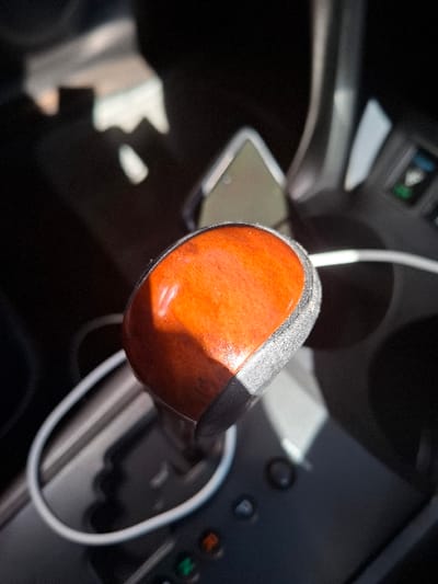 The image shows the gear shifter of a car, likely located in a vehicle in Gitaru, Kenya, at the coordinates provided. The gear shifter is in the park position, with the shifter knob visible. The knob is brown and appears to be made of wood. It is surrounded by the black interior of the vehicle. The shifter base is silver with a black outline. A white cord is wrapped around the shifter base, suggesting it is connected to a device. The image also shows a part of the car's console, with several buttons and a control panel in the background. The lighting is bright, suggesting it is daytime and the car is parked. No people are present in the image.