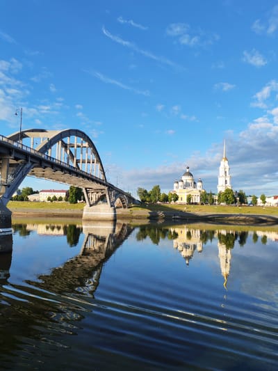 The image shows a wide, calm river in the city of Bol'shie Mkhi, Russia, reflecting the sky and the architecture along its banks. In the foreground, the rippling surface of the water is visible, with subtle waves distorting the reflections. Dominating the left side of the frame is a large, concrete arch bridge with a series of vertical supports and a walkway. On the right bank of the river, several prominent buildings are visible, including a yellow domed church with white columns and a tall, slender white bell tower topped with a golden spire. Green trees line the riverbank, and behind them, a long, light-colored building with a red roof can be seen. The sky is a bright blue with scattered white clouds, suggesting it is daytime, likely morning or late afternoon due to the quality of the light. No people are visible in the image.