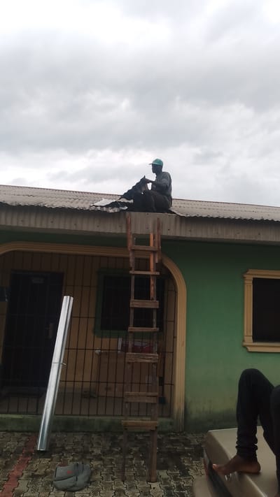 The photograph, taken in what appears to be Asubi, Nigeria, presents a single-story building under an overcast sky. The building's exterior walls are painted in a combination of green and tan, with security bars covering the window and door. A man is seated on the corrugated iron roof, seemingly working with dark material. A weathered wooden ladder leans against the building, providing access to the roof. At ground level, a long, silver metal pipe rests against the security bars. Adjacent to the pipe, a grey duffel bag lies on the paved surface. In the foreground, part of a car's trunk is visible, with a person seated on it; only the lower legs and bare feet are captured.