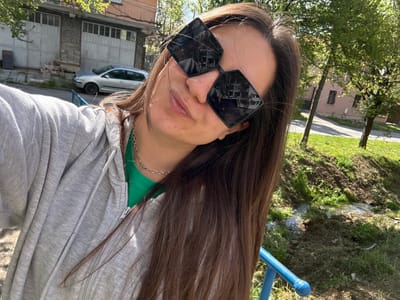 The image captures a close-up, upward-angled portrait of a person, likely female, with long, dark brown hair. She is wearing large, dark, geometric-shaped sunglasses that cover most of her eyes, and a relaxed expression is suggested by the slightly parted lips. Her attire consists of a bright green top, visible under an unzipped light gray hooded jacket. A delicate silver chain necklace adorns her neck.

The scene is set outdoors in bright daylight, indicating a sunny day in Radomir, Bulgaria. In the background to the person's left, there is a natural area of grass and dirt, with a blue metal pole suggesting a possible fence or playground structure. To the person's right and in the mid-ground, several buildings of varying architectural styles are visible, alongside a white parked car. Trees with vibrant green foliage also contribute to the outdoor setting, and a paved road or path can be seen winding through the background. There is no visible text in the image. The overall impression is one of a casual moment captured in an urban or semi-urban outdoor environment.