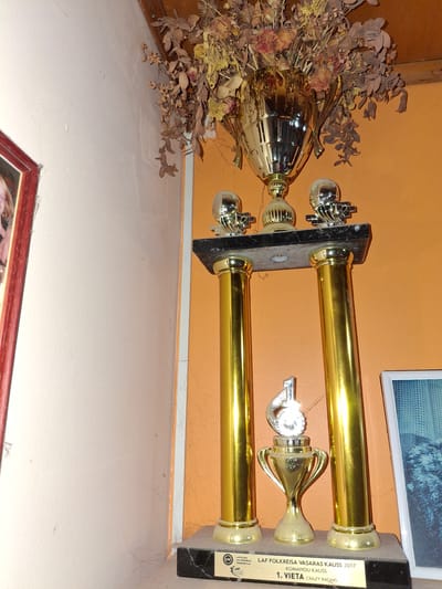 The media file depicts a collection of trophies displayed on an orange-colored wall, with a cream-colored wall below. The trophies are predominantly gold, featuring cylindrical bases and intricate decorative elements. One trophy has a large, globe-like component and is adorned with dried flowers. Another trophy, positioned lower, features a smaller cup-like structure and a silver whistle. A prominent trophy on the right side has a black base with visible text.
The visible text on the black base reads: "LAF FOLKREISA VASARAS KAUSS 2017 KOMANDU KAUSS 1. VIETA CRAZY RACING" and "LATVIJAS AUTOMOBILU FEDERĀCIJA". This suggests the trophies are related to a 2017 summer folk race event, specifically a team competition titled "Crazy Racing," where a team achieved first place. The presence of the Latvian Automobile Federation (LAF) logo further solidifies this context.
The scene appears to be indoors, possibly a home or a trophy display area, given the setting and the inclusion of dried flowers. The lighting suggests it might be daytime, but the artificial light also plays a role. The overall impression is one of achievement and recognition, likely in the context of motorsport or a related racing event.