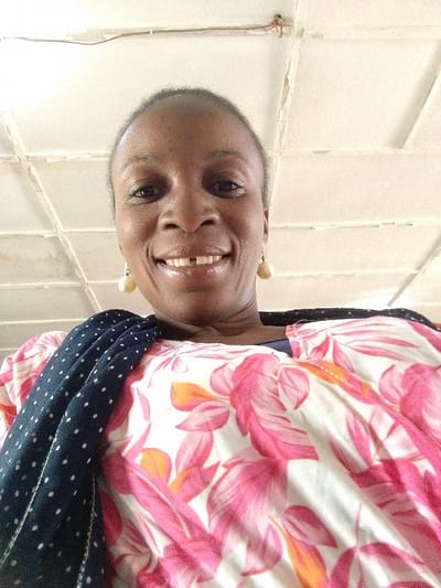 An adult female is depicted in a low-angle close-up, smiling directly at the camera. She has short dark hair, dark eyes, and a visible gap between her two front upper teeth. She wears light-colored, spherical drop earrings and a loose-fitting garment with a white background featuring a repeating pattern of pink and orange tropical leaves. A dark blue fabric with white polka dots is draped over her left shoulder. The background consists of a light-colored, possibly off-white, tiled ceiling with visible grid lines and some discolored or damaged sections, including what appears to be exposed wiring or a fixture near the top left corner. The scene is located indoors, within the city of Barnawa, Nigeria.