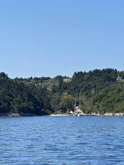 The image depicts a coastal scene on the island of Antipaxos, Greece. The foreground is dominated by the blue, rippling water of the Ionian Sea, reflecting the clear blue sky above. The middle ground showcases a lush, green hillside covered in dense evergreen trees and some olive trees. Several houses with red-tiled roofs are visible nestled amongst the trees on the upper slopes of the hill. At the base of the hill, a small, sandy cove can be seen, with a few boats anchored in the shallow water. Two white sailboats are prominently moored near the shore. The overall impression is one of a serene, Mediterranean landscape.