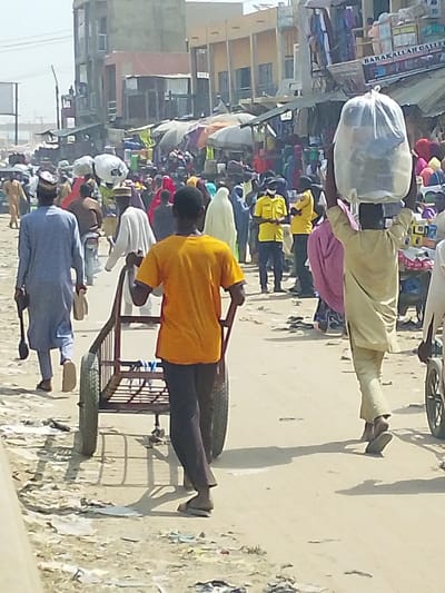 The image depicts a bustling street scene within a market area in Fagge D1, Nigeria, under bright daylight. In the immediate foreground, facing away from the viewer, a male individual wearing an orange t-shirt and dark trousers pushes an empty, reddish-brown, two-wheeled handcart. To his right, another male individual, also facing away, wears a light tan traditional garment and carries a large, clear plastic bag filled with items balanced on his head. To the left of the cart-pusher, a male in a light blue traditional garment walks away, holding a small light-colored object in his left hand.
The midground is densely populated with numerous pedestrians moving in various directions, many carrying goods, some on their heads. Market stalls with assorted merchandise and colorful awnings or umbrellas line both sides of the street. Notable figures include two individuals in bright yellow shirts, dark trousers, and face masks standing near the center-right. Several women are visible, some wearing light-colored hijabs or full veils, and one in a purple garment.
In the background, multi-story commercial buildings are visible, one featuring a sign with "BARAKALLAH GALLERY" prominently displayed. The ground is an unpaved, dusty surface scattered with debris and litter. Strong sunlight casts distinct shadows, indicating a clear, bright sky.