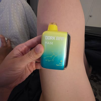 A hand holds a "DORK BAR PULSE" vape device, which is yellow to blue gradient with constellations and the text "Sour Apple Ice" printed on it. The device is held against a person's thigh, which is bare and light-skinned. In the background, a white and gray patterned blanket is visible. The environment appears to be indoors, possibly a bedroom. No specific location in Portland, United States of America can be determined from the image.