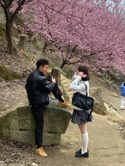 A man in a black jacket and pants with tan boots sits on a rock, photographing a young woman standing on a higher rock. She wears a white blouse, gray pleated skirt, white socks, and black shoes, accessorized with a black shoulder bag and a pink hair accessory. A blooming tree with pink flowers cascades into the frame to the left of the pair. Other individuals and rocks are visible in the background.