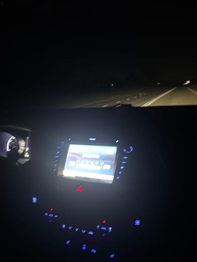 The image shows the interior of a car at night, looking forward through the windshield and at the dashboard. The dashboard is illuminated by the car's interior lights and the glow of the infotainment screen. The screen displays what appears to be radio information, with the frequency "101.50 MHz" visible. Buttons and indicators on the dashboard are also lit, some with blue and red lights. The road ahead is visible through the windshield, with lane markers faintly illuminated. The surrounding environment is dark, indicating a rural or sparsely populated area, possibly on a highway. The image does not contain information that would allow for specific location identification within Mokryi Batai, Russia.