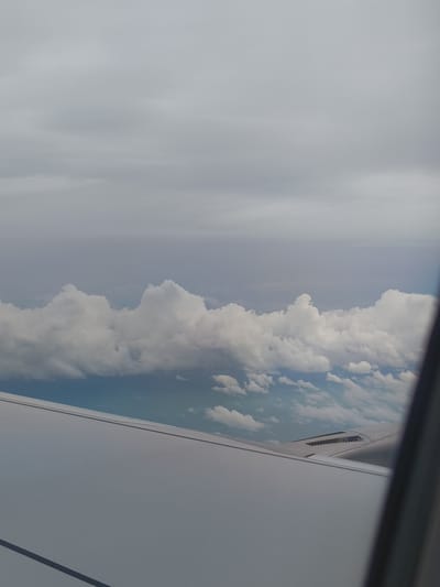 The image is a photograph taken from an airplane window, looking out at a cloudy sky. The lower portion of the image shows the wing of the airplane, which is light gray and smooth. Above the wing, there are layers of clouds. The clouds closest to the wing are white and fluffy, appearing to be cumulus clouds. Further up, the clouds become more diffused and gray, suggesting a layer of stratus clouds or an overcast sky. The overall lighting is muted, indicating it might be daytime with heavy cloud cover. There are no people or other identifiable objects visible in the scene other than the airplane wing and the sky. The image does not depict any specific location in the city of Giồng Kè, Vietnam.