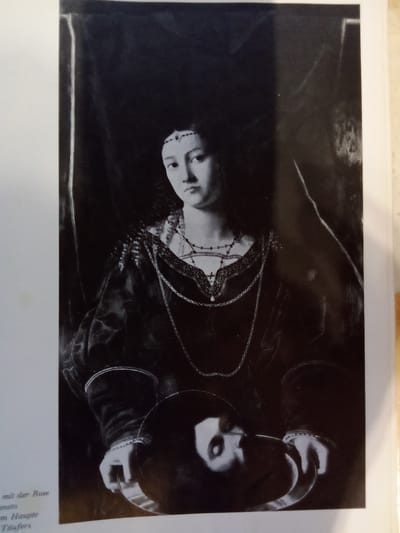 This is a black and white photograph of a painting. The painting depicts a woman in elaborate clothing, wearing a jeweled headband and multiple necklaces. She is holding a silver platter in her hands, on which rests the severed head of a man. The woman's expression is calm and composed. The background is dark and indistinct, with some suggestion of drapery. The lighting is dramatic, highlighting the faces of the woman and the man.

The image is a reproduction, with some text visible in the lower left corner, partially cut off. The legible text includes German words such as "mit der Rose," "im Haupte," and "Täufers," which translate to "with the rose," "on the head," and "of the baptizer" respectively. These fragments suggest the painting might be a depiction of Salome with the head of John the Baptist, or a similar subject. The overall mood of the painting is somber and dramatic.