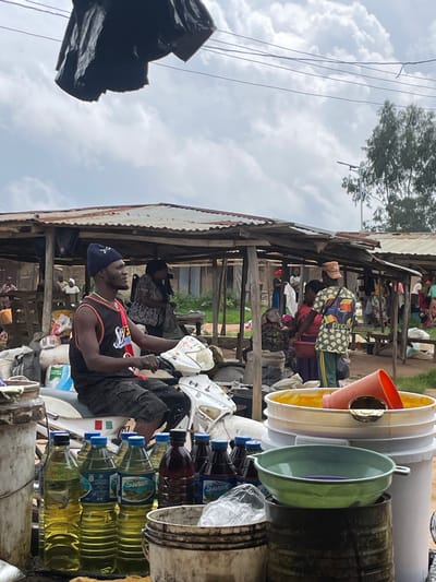 The image depicts an outdoor market scene, likely in Wang, Nigeria. In the foreground, a man wearing a black beanie and a sleeveless basketball jersey is seated on a white motorcycle. He is looking to his right and appears to be interacting with something out of frame or waiting. Next to the motorcycle are several large plastic bottles filled with yellow liquid, possibly oil or fuel, and some dark liquid in bottles. A large white bucket topped with a yellow substance and an orange funnel, and a green bowl are also visible in the foreground.
Behind the motorcycle, a series of makeshift stalls with corrugated metal roofs are set up. Several other individuals are present in the background, some are seen within the stalls, and others are walking through the market. The overall environment appears to be a busy, informal market with various goods and people. The sky is cloudy.