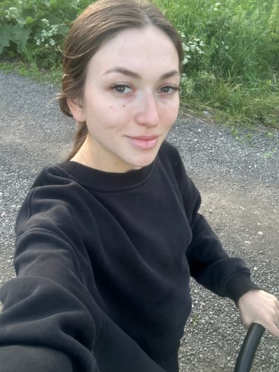A young woman with brown hair and fair skin is taking a selfie outdoors. She is wearing a black crewneck sweatshirt. The background consists of a gravel path and lush green foliage. The overall lighting suggests it is daytime, likely late afternoon or early evening, with soft natural light. The location is Estachi, Russia.